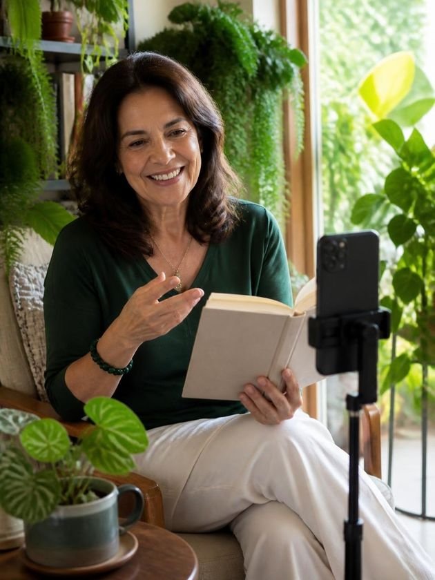 Criadora de conteúdo sobre livros e plantas fala para o celular num tripé, com um livro aberto na mão, num canto de leitura cheio de plantas.