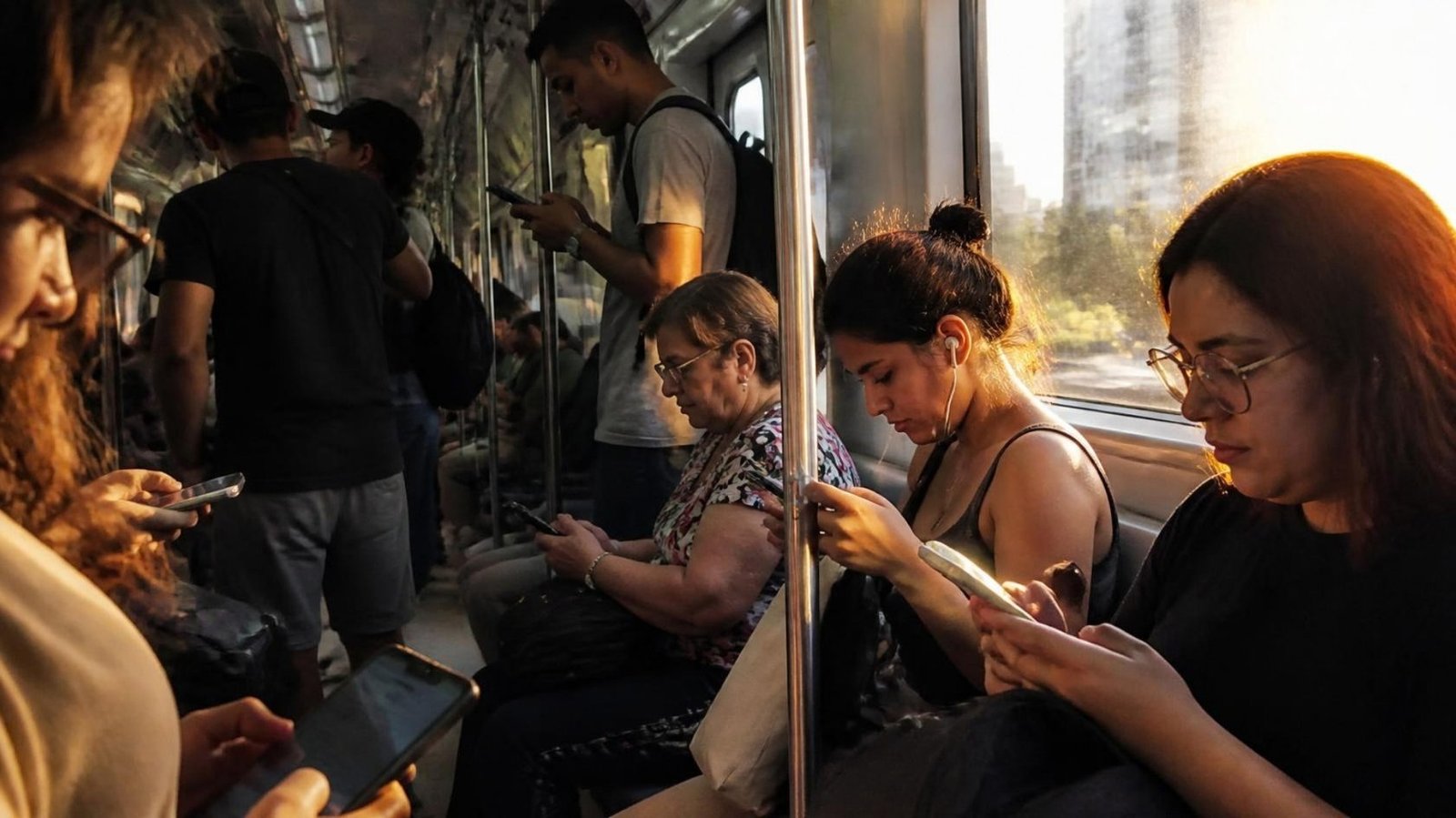 Passageiros num vagão de metrô no fim da tarde, cada um olhando o próprio celular, com a luz dourada entrando pela janela.